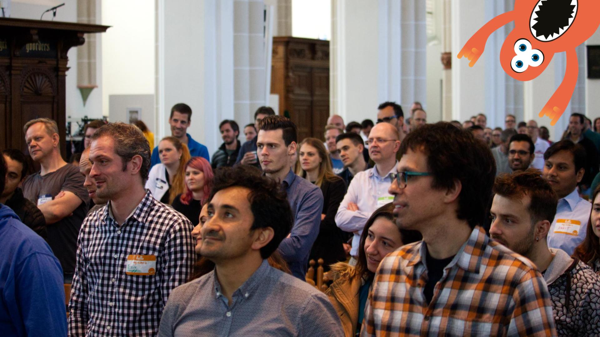 A large group of people stood up at a TestBash conference. There is an orange monster character peeking in at the top right of the frame.