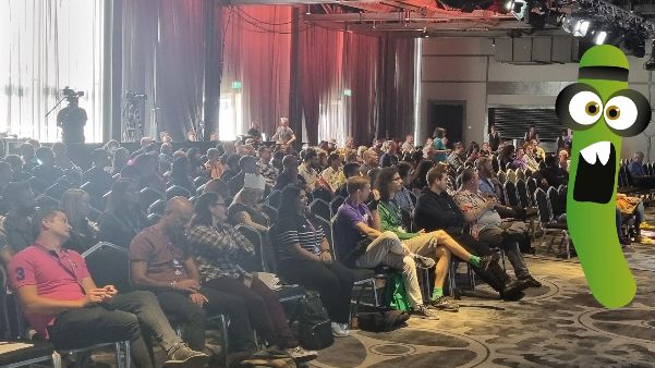 A large group of people sat in a conference space. There is a green monster character near the stage which is out of shot.