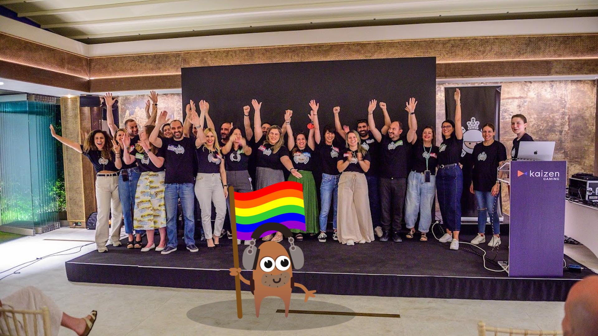 A large group of people on a stage with their arms raised. There is a monster character in the foreground holding a rainbow flag. Credit. MoT Athens.