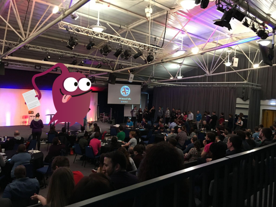 A crowd of people in an auditorium. There is a queue of people waiting to go on stage. There is a magenta monster character on stage. 