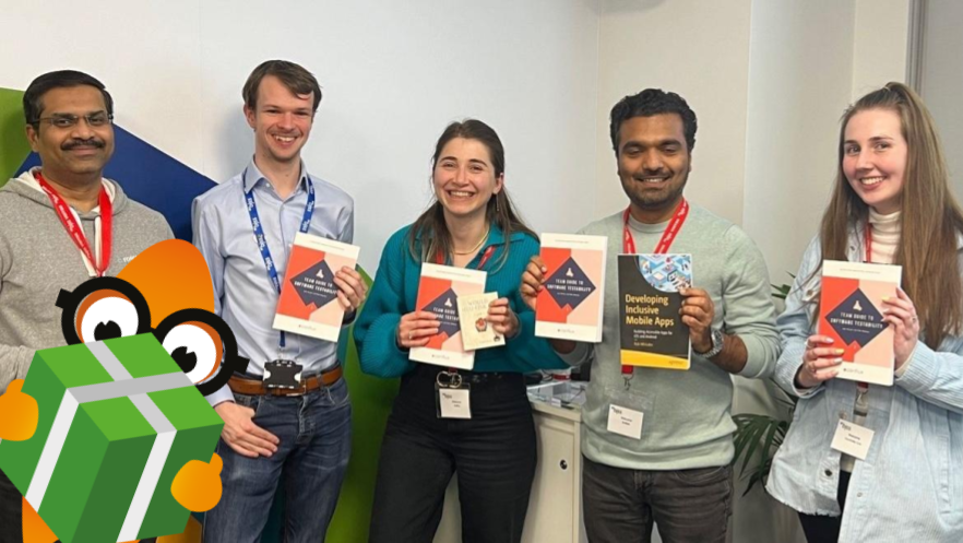 Five people stood next to each other smiling and holding books. An orange monster with a green gift box is photobombing the photo. Credit: Leeds MoT Meetup