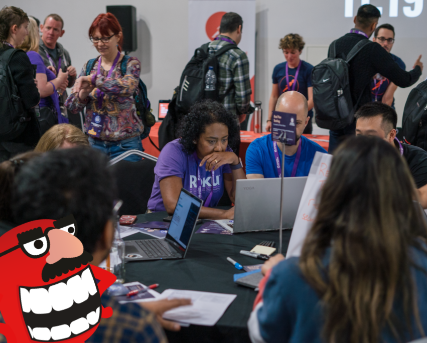 A group of people gathered around a table. There are people standing and discussing things in the background. There is a red monster character that is wearing a disguise.
