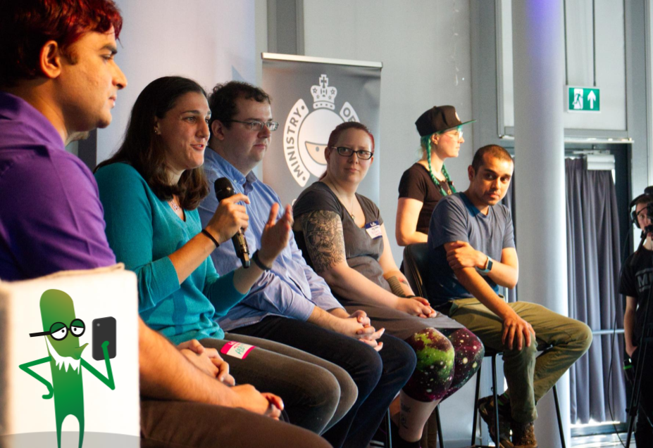 A panel of men and women sat on high rise chairs on stage speaking at a conference. There is a green monster character holding a phone in the foreground.