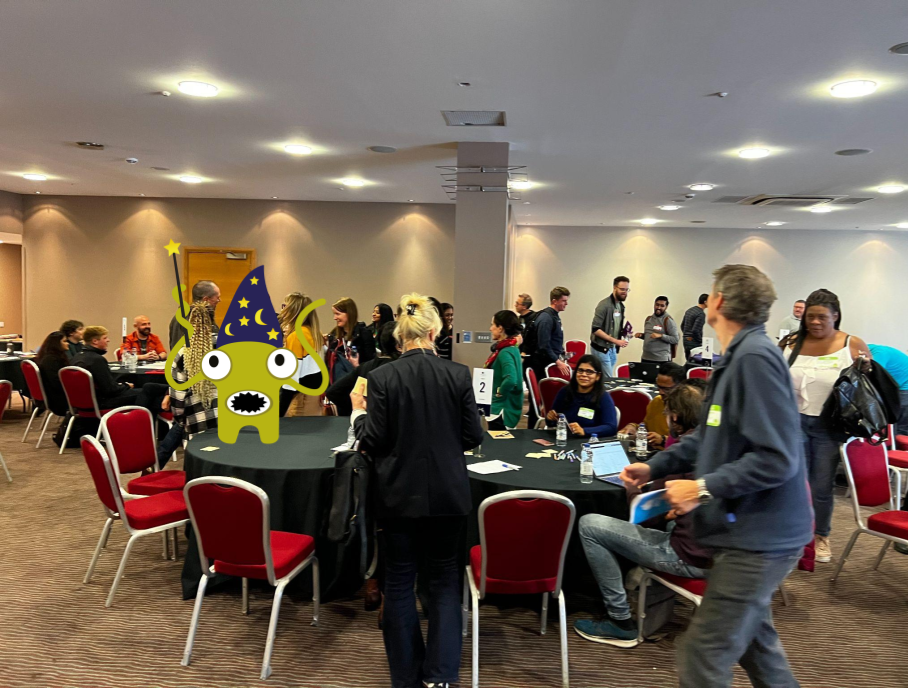 People gathered in a room for workshops. There are circular tables. There is a green monster character on a table.