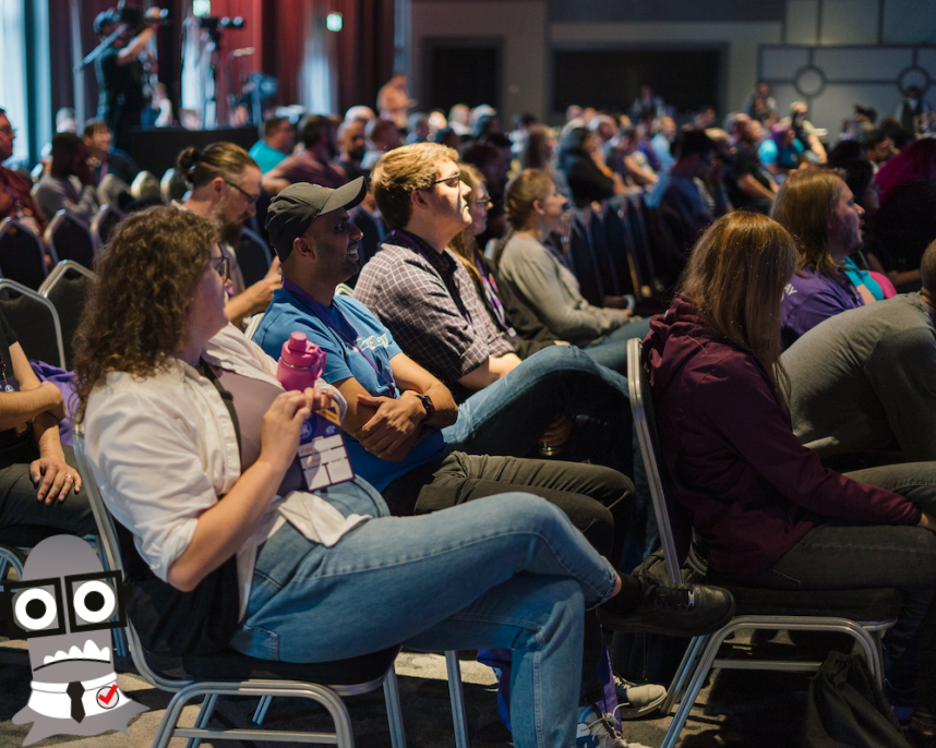 A group of people sat listening to a talk in a conference hall. There is a monster character hiding near a chair.