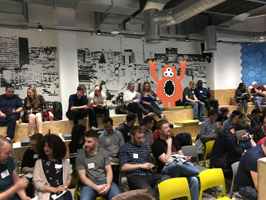 People gathered in a small lecture theatre setting. People are sat paired up running an exercise. There is an orange monster character with their hands in the air making the rock sign with their fingers.