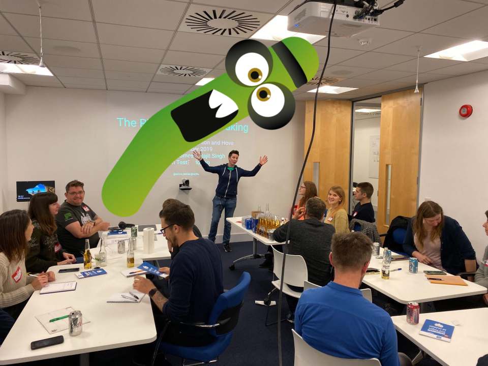 A small group of people in a workshop. There is a person with their arms wide at the front. A green monster character is photobombing. 