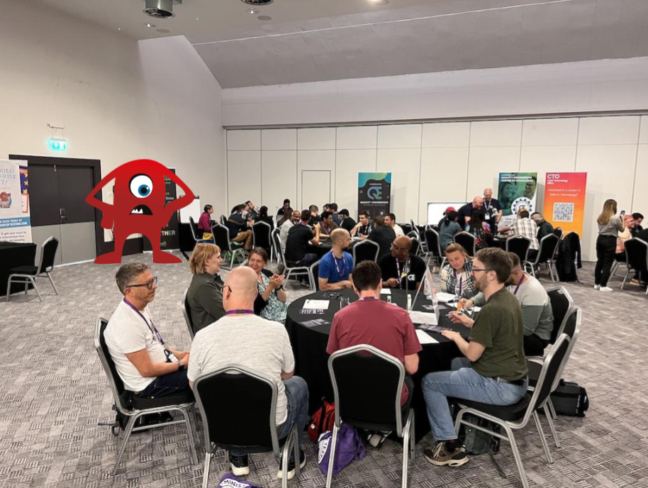 Groups of people gathered around tables working on exercises during a conference workshop. There is a one-eyed red monster character next to one of the tables.