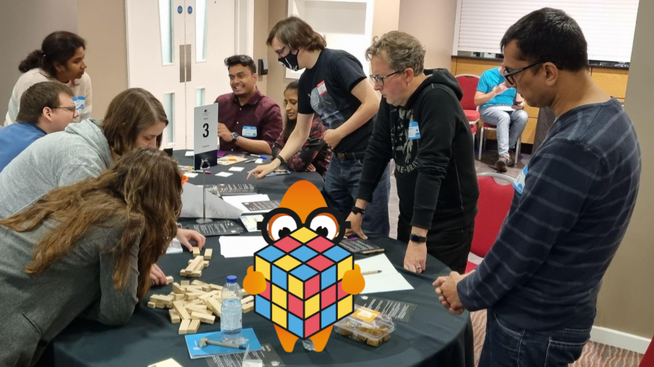 People gathered around tables. They are running an exercise at a testing event. There is an orange monster character in the foreground. It is holding a cubed puzzle object.