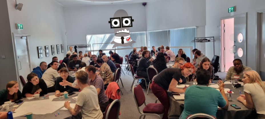Several people sat at several tables. There is a grey monster character at the front of the room.